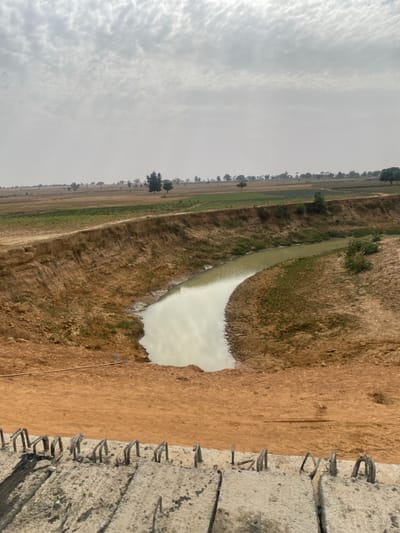 The image depicts a landscape in Wuciciri, Nigeria, characterized by a narrow waterway flanked by earthen banks under a cloudy sky. In the immediate foreground, the top surface of a concrete structure, possibly a foundation or part of a bridge, is visible. This structure consists of multiple grey, rectangular concrete blocks with exposed steel rebar loops projecting upwards from their surface. Directly behind this, a reddish-brown earth expanse slopes downwards towards a winding, light brown or greenish-beige body of water, approximately 5-7 meters wide, reflecting the cloudy sky. The water's banks are steep, unpaved earthen slopes, displaying varying textures and some sparse green vegetation. Beyond the waterway, a flat, expansive landscape extends to the horizon. This midground and background consist of large tracts of agricultural land, showing patches of tilled brown earth and cultivated green fields. Scattered small to medium-sized trees are distributed across this distant terrain. The sky is overcast with diffuse grey and white clouds, providing consistent ambient light. No individuals, animals, or explicit actions are visible within the frame.