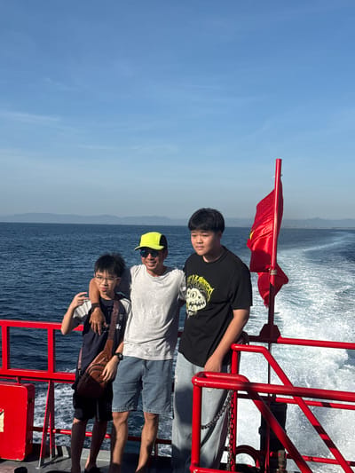 Three males are positioned on the deck of a red-railed boat sailing on a body of water under a clear blue sky. The male on the far left, a young boy with black hair and glasses, is wearing a dark t-shirt, shorts, and a brown sling bag. He is making a peace sign with his left hand. The central male, an adult with black hair and a neon yellow baseball cap, is wearing a grey t-shirt and denim shorts. He has his arm around the boy on his left. The male on the far right, a young boy with black hair, is wearing a black t-shirt with a graphic design and light-wash denim pants. He is looking towards the right. A red flag with a yellow star is attached to a pole behind the group, partially obscuring the view of the water and a wake trailing behind the boat. The background shows a distant coastline with mountains. The scene suggests they are on a boat trip, possibly near An Hải, Vietnam, given the presence of a Vietnamese flag.