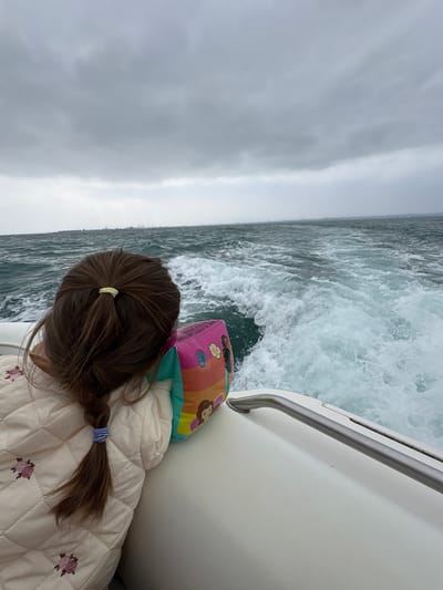 A young girl with brown hair in a braid is riding on a boat. She is wearing a quilted jacket with floral embroidery. She is holding a pink floatation device with images of cartoon characters. The boat is moving through choppy, dark blue water, leaving a white wake. The sky is overcast and grey. In the distance, there is a hazy shoreline with industrial structures, possibly indicating a port or city. The scene is likely a coastal area near La Punta, Peru, known for its maritime activities and proximity to Lima.