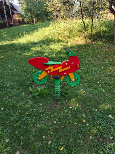 A red and green spring-mounted toy motorcycle is positioned on a grassy lawn in a wooded area. The motorcycle has yellow zigzag decorations on its sides and green wheels. In the background, a rustic wooden building with a gray roof is visible among trees. The ground is covered in green grass with some scattered fallen leaves. The lighting suggests it is daytime.