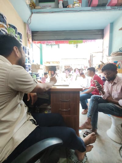 An interior shot captures a retail space in the city of Balla, Bangladesh, opening onto a bright, busy street scene. In the foreground, a male individual, observed from behind, is seated in a black office chair at a wooden desk. He wears a tan short-sleeved shirt and dark trousers, with his right forearm resting on the desk near an open notebook. His feet are bare. To the right of the desk, another male, wearing a light pink shirt and blue jeans, sits on a white cylindrical stool, engrossed in a smartphone held in both hands. His feet, clad in flip-flops, rest on the floor. A third male, in a red shirt and blue jeans, is partially visible behind the desk, looking towards the right. The shop's left wall displays shelves stacked with multiple paint cans, some bearing "Dulux" branding. The shop interior features light blue walls and a tiled floor. Above the open storefront, colorful decorative streamers are visible. The exterior reveals a bustling street with several pedestrians and distant structures under bright daylight.