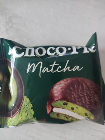 The image shows the packaging for a matcha-flavored Choco Pie. The packaging is predominantly dark green with white text that reads "Choco Pie" in a stylized font, followed by "Matcha" in a cursive font. A depiction of the matcha Choco Pie, showing its chocolate coating, green cake layers, and white filling, is visible on the right side of the packaging. On the left side, there's an abstract depiction of matcha powder and a portion of a matcha drink. The packaging is lying on a light-colored, textured surface that could be a table or countertop. There are no people or discernable elements that would allow for the identification of a specific location in Gorno-Altaysk, Russia. The focus is entirely on the product packaging.