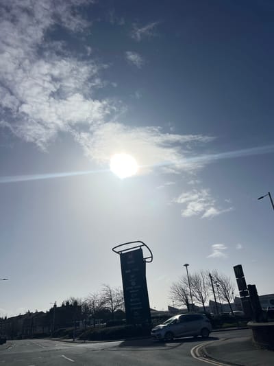 The image depicts an outdoor, daytime scene at the Meols Cop Retail Park in Southport, United Kingdom. The sky is bright blue with scattered white clouds, and the sun is visible as an intense, overexposed white circle with lens flare, positioned centrally in the upper frame.
In the midground, a large, dark-colored vertical sign stands prominently, displaying "M PARK Southport Retail & Leisure Park" at its top. Below this, a list of businesses is visible, including ASDA, B&M, NEXT, Sports Direct, Pets at Home, M&S Food, Iceland, Curry's PC World, McDonald's, and Costa Coffee. To the right of this sign, a smaller, dark sign with the McDonald's logo is partially visible.
An asphalt road with a slight curve is visible in the lower foreground, featuring a double yellow line near the right edge. A silver four-door crossover vehicle is positioned on this road, facing right, in front of the retail park entrance. Bare deciduous trees are scattered behind the main sign and throughout the midground, indicating a cooler season. Several streetlights are also present.
In the far background on the left, a row of residential terraced houses with dark roofs is visible against the horizon. No individuals are discernible in the scene.