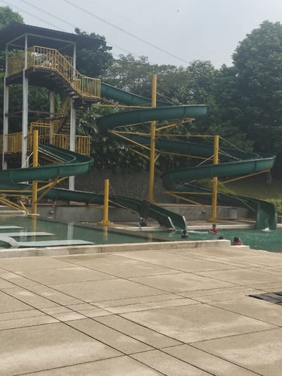 This is a wide-angle, eye-level, outdoor, daytime photograph of a water slide at a swimming pool facility in Gunung Putri, Indonesia. The water slide is green and yellow, with a spiral design. It is supported by yellow metal beams. There are stairs leading up to the slide. Several people are visible in the swimming pool, which is filled with turquoise water. The pool area is paved with light-colored tiles. Trees and a stone wall are visible in the background. The sky is overcast.