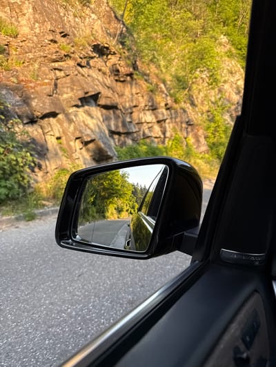 This media file captures a view from inside a car, looking out the side window towards the right. The primary focus is the side-view mirror, which reflects a road winding through lush green trees and foliage. The angle of the shot suggests the car is in motion, likely on a scenic route.
To the left of the mirror, a rocky cliff face is visible, bathed in warm sunlight, indicating it's likely late afternoon or early morning. Small green bushes and grasses grow at the base of the cliff and in crevices. The road surface, seen through the mirror and to the right of the car, appears to be asphalt. The sky is bright, suggesting clear weather.
Inside the car, part of the door panel is visible, including a speaker grille with the "Harman Kardon" logo. The interior is dark and out of focus, drawing attention to the external scenery. The overall impression is one of a pleasant drive through a natural landscape. No people are visible. The location is identified as Taran, Bulgaria.