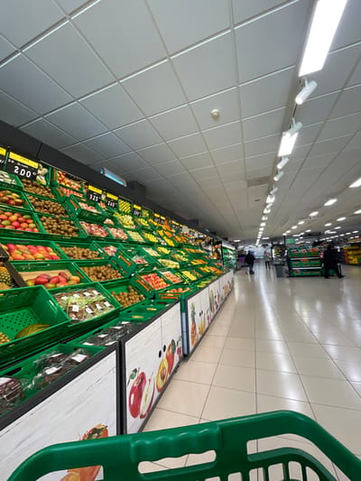 The scene depicts the interior of a supermarket, specifically the produce section, located in Altafulla, Spain. The perspective is from behind a green shopping cart, whose handle is visible in the lower foreground. To the left, a long, multi-tiered display of fresh fruits and vegetables is arranged in numerous green plastic crates. Visible produce includes mangoes, kiwi fruit, apples, pineapples, and other assorted items. Black price signs above the produce indicate costs in Euros, such as "2,00€" and "3,25€", with some labels like "PIÑA" (pineapple) and "KIWI" discernible. The base of the produce display features decorative white panels with colorful illustrations of various fruits.
A wide, light-tiled aisle extends into the background, illuminated by overhead fluorescent panel lights and white cylindrical track spotlights set into a white grid-tile ceiling. A circular smoke detector or sprinkler head is visible on the ceiling. In the middle to far background of the aisle, approximately four individuals are present. One person in dark clothing is seated on what appears to be a bench or mobility scooter on the right side of the aisle. Another person is walking with a small cart, and two other figures are partially visible further down the aisle, all in varying states of motion or observation within the commercial setting.