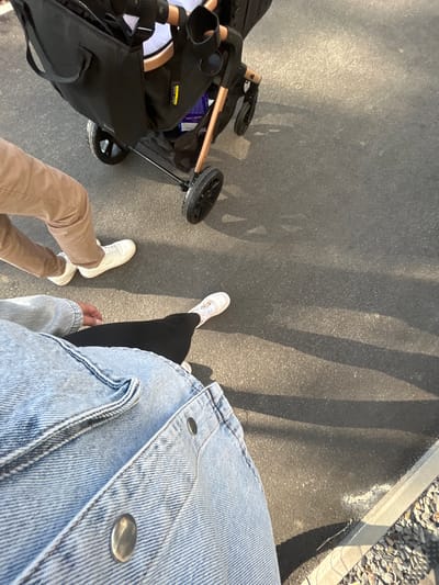 Two individuals are walking on an asphalt surface. The person in the foreground is wearing a light blue denim jacket and black leggings, with one foot visible, wearing a white sneaker with pink accents. They are in the process of walking forward. Behind them, another person is walking and pushing a black stroller. This individual is wearing tan pants and white sneakers. The stroller has a wooden handle and black wheels. The ground is a dark asphalt surface, with shadows cast by the stroller and possibly other unseen objects. In the lower right corner, a concrete curb is visible, adjacent to a gravel-covered area. The scene appears to be outdoors, possibly on a sidewalk or a paved path. The location is identified as Waldbröl, Germany.