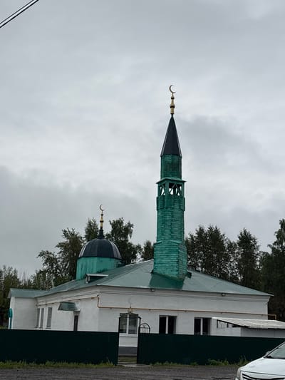 The image shows a mosque in Krasnokamsk, Russia. The mosque is a white building with a green roof and a tall, green minaret topped with a crescent moon. There is also a smaller dome with a crescent moon. The mosque is surrounded by trees and a dark green fence. The sky is overcast.