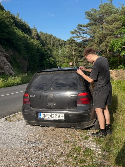 In Pechinska, Bulgaria, a young man with curly hair and wearing a dark t-shirt and shorts is standing next to the rear of a dark-colored Volkswagen Golf. He is using his hands to adjust something on the top edge of the car's rear window. The car is parked on the side of a road, with a gravel shoulder and grassy embankment. The background features a steep, tree-covered hillside under a clear blue sky. A traffic sign is visible on the hillside behind the car. The license plate of the car reads "CM 9422 AH" with Bulgarian markings.