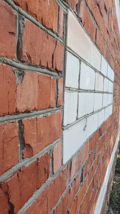 A close-up, angled shot showcases a weathered brick wall in Ust-Labinsk, Russia. The wall features a section of white bricks integrated into the red brickwork, creating a distinct rectangular pattern. The red bricks exhibit signs of age, with visible cracks and imperfections. The mortar between the bricks is grey and appears to be aged. The lighting suggests an outdoor environment, possibly under natural sunlight. The background is blurred, but hints of green grass and earth are discernible, indicating an exterior setting. No people or specific actions are depicted.