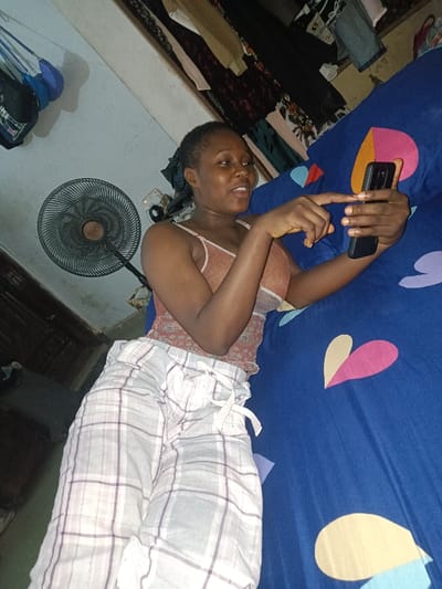 A woman lies on a bed with blue heart-patterned sheets, looking at a smartphone in her hand. She's wearing pajamas with a white and grey plaid pattern and a spaghetti-strap top. A standing fan is positioned next to the bed, and a closet filled with clothes is visible in the background. The room appears to be dimly lit.