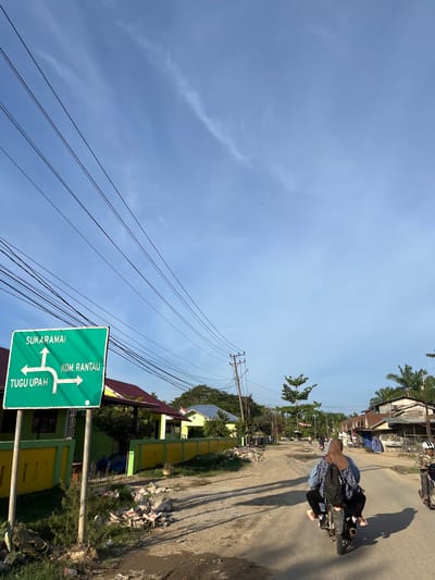 The image depicts a street scene in Alurcucur, Indonesia, under a bright, clear blue sky with scattered wispy clouds. A prominent green directional sign is visible on the left, indicating "SUKARAMAI" with an upward arrow, "TUGU UPAH" with a leftward arrow, and "KOM. RANTAU" with a rightward arrow. The sign is mounted on two metal poles. To the right of the sign, a dirt road stretches into the distance, lined with houses and shops on both sides. Several power lines are strung across the sky.
In the foreground, a dirt road is visible, with debris and small piles of rubble scattered along its edge, particularly near the sign. The road surface appears uneven and dusty.
Several people are present in the scene. Two individuals are riding a motorcycle in the middle of the road, facing away from the camera. The person in the back is wearing a hijab. Further down the road, two more motorcycles are visible with riders, and another person appears to be walking.
The buildings lining the street are varied in style and condition. Some appear to be houses with brightly colored walls (yellow and green are noticeable) and red or blue roofs. Others seem to be more modest shops or residences with corrugated metal roofs. Vegetation, including trees and palm trees, is present throughout the scene, adding to the tropical atmosphere. The lighting suggests it is daytime, likely late afternoon given the long shadows cast by the power lines and buildings.