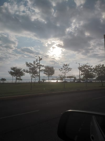 The image captures a view of the Encarnación waterfront in Paraguay. The foreground shows a dark asphalt road with a white line marking its edge, and the driver's side rearview mirror of a vehicle. The middle ground features a well-maintained grassy area bordered by a yellow curb. Beyond the grass, a walkway or promenade runs parallel to a body of water, likely the Paraná River, which is visible in the background.
A row of trees, some with sparse foliage, are planted along the walkway. Lampposts are interspersed among the trees, and a few figures are visible in silhouette, suggesting people are walking or sitting near the water. Several cars are parked or driving along a road adjacent to the walkway. The water stretches out to the horizon, where it meets a hazy sky.
The sky is dramatic, filled with a mix of dark, heavy clouds and brighter patches where the sun is attempting to break through. The sun is positioned high and slightly to the right of center, its rays diffused by the clouds, creating a soft, atmospheric light. The overall impression is of a cloudy, possibly overcast day, with the sun providing a gentle illumination. The scene is tranquil, conveying a sense of a public park or recreational area along a waterfront.
