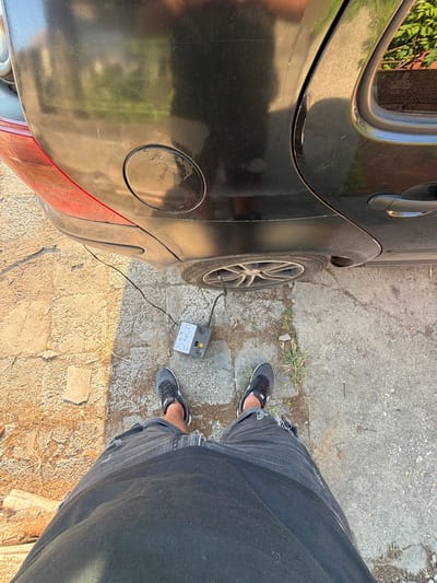 The image captures a first-person, downward-looking perspective of an individual using an air compressor to inflate a car tire in Kalipetrovo, Bulgaria. The scene is set outdoors on a sunny day, with bright, direct sunlight creating distinct shadows.

The subject's lower body is visible, wearing dark jeans, possibly with some distressing or rips, and dark athletic shoes with white accents. They are standing on a ground surface that appears to be a mix of cracked asphalt, small stones, and patches of dry grass or weeds.

To the left, a black car, likely a hatchback, is partially visible. The rear passenger door, the rear wheel, and the fuel cap are prominent. The car's body shows some dirt and minor scratches. A portable air compressor, primarily grey with yellow/orange accents, is positioned on the ground next to the car's rear wheel. A black hose extends from the compressor, connecting directly to the valve of the car's tire, indicating the inflation process is underway.

The air compressor features a gauge and several symbolic icons on its surface, depicting various items that can be inflated, such as car tires, bicycle tires, and sports balls. While specific text is unreadable, these symbols likely correspond to operational instructions or pressure settings. The overall impression is one of practical car maintenance taking place in a rustic, outdoor environment.