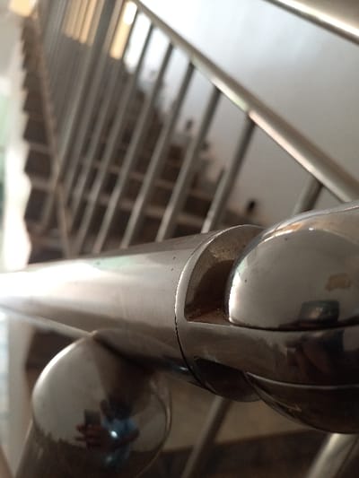 This image depicts a close-up, upward-angled view of a metallic staircase handrail and balusters within a building in Oturkpo, Nigeria. The foreground is dominated by a polished, cylindrical metal handrail. A decorative spherical metal element is connected to the handrail via a visible joint, which shows an accumulation of light brown particulate matter. On a lower, spherical portion of the railing, a blurred reflection of a human hand and arm, wearing a blue garment, is visible. The staircase extends into the blurred background, featuring multiple vertical metal balusters and dark-colored steps. A light-colored wall runs along the right side of the staircase. No other individuals or specific actions are discernible. The environment is an interior architectural space.