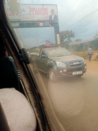 A dark grey Isuzu D-Max pickup truck is driving on a dirt road, kicking up dust. The truck has a black roll cage and a red object on top, possibly part of a structure for carrying items. The license plate reads "KDB 249E". In the foreground, viewed through a window, is the interior of a vehicle, with a person's shoulder and head visible in the passenger seat.
Above the road, a large billboard is visible. The billboard has text that reads "US AL WELCOME TO THE FUTURE". A man in a suit is pictured on the billboard. Another smaller sign below the billboard says "WELCOME".
To the right of the road, a man in a plaid shirt and dark pants stands looking towards the truck. The setting appears to be a rural or semi-urban area in Kenya, given the style of the billboard, the vehicle, and the presence of a dirt road. The sky is overcast, suggesting a cloudy or hazy day. The overall impression is of a dusty, possibly hot, environment.
---
**Note:** The initial request included a "Location context: Hospital ward, Kenya", but the visual content of the image does not depict a hospital ward. The description provided is based solely on the visual elements present in the image.