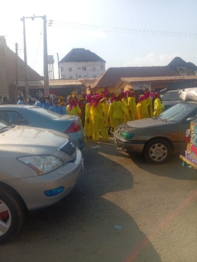 The image depicts a street scene in Ozuri, Nigeria, under daylight conditions. In the mid-ground, a large group of individuals is visible, primarily adorned in bright yellow long-sleeved garments and headwear featuring prominent pink feather-like decorations, suggesting a procession or organized gathering. Behind them, a smaller group in blue attire is partially discernible.
The foreground contains parked vehicles: a silver sedan occupies the left, displaying blue-tinted fog lights and a chrome grille. To its right, a dark green or grey sedan is visible. On the far right, part of a colorfully painted commercial vehicle, possibly a truck or minivan, is present.
The background features various structures, including a prominent multi-story light-colored building with a dark hip roof. Smaller single-story buildings with brown roofs are also visible. On the far left, a tall brick wall runs alongside the road, behind which utility poles with power lines and a metal water tower structure are present. The asphalt road surface is visible, with shadows cast to the left, indicating strong sunlight from the right. The sky is clear and light blue.