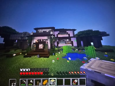A Minecraft scene shows a two-story house with white walls and a dark roof beside a small pond. The house is decorated with foliage. The player's health bar indicates they have 9 full hearts. The inventory bar shows various items, including bread, arrows, and a pickaxe. The sky is dark, suggesting it is nighttime in the game. A wooden structure with log-like textures is visible to the left of the screen, possibly a table or building component.