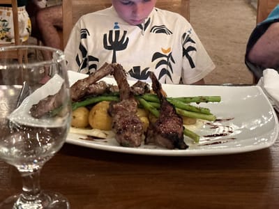 The image shows a plate of lamb chops with asparagus and small potatoes, drizzled with balsamic glaze, on a white rectangular plate. A partially filled water glass is in the foreground, slightly blurred. A child wearing a white T-shirt with black geometric designs is seated behind the plate. Other people are seated to the left and right of the child, out of focus. The scene appears to be a restaurant in Tamarindo, Costa Rica. The plate rests on a dark wooden table.