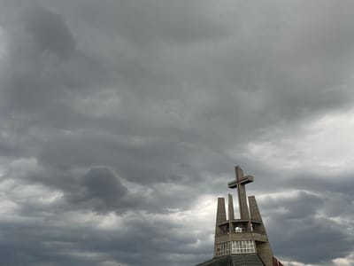 The image shows the upper part of a modern concrete church tower with a cross at its apex, set against a dramatic, cloudy sky. The tower features a unique, angular design with open sections that reveal bells and balconies. Large windows are visible on the lower portion of the tower. The sky is filled with dark, heavy clouds, suggesting an overcast or stormy weather. This is the tower of the Church of the Peace in Świdnica, Poland, a UNESCO World Heritage site. No people are visible in the image.