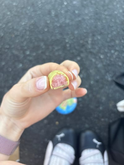 A close-up shot shows a person's hand holding a bite-sized food item. The food item has a light yellow outer shell with a pink, crumbly interior. The hand has neatly manicured nails with a French manicure. In the background, out of focus, are a pair of dark shoes with white laces, and a small, colorful globe toy. The scene appears to be outdoors, with a dark, textured ground that looks like asphalt or pavement. The lighting suggests it is daytime. The overall impression is casual, perhaps a snack being enjoyed outdoors. The location context provided is Erlenbach am Main, Germany. There is no visible text in the image.