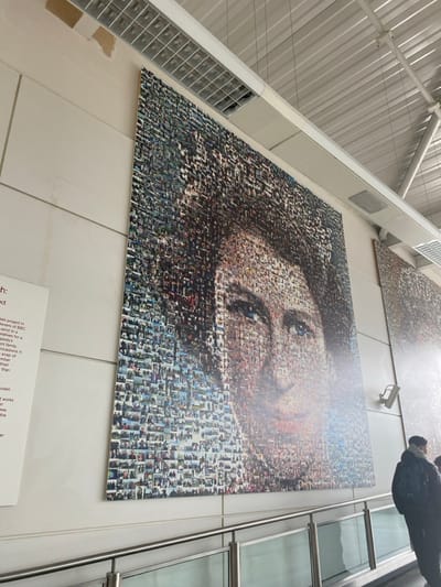 A large-scale mosaic artwork depicting the face of a woman is prominently displayed on a light-colored, possibly white, panelled wall. The artwork, which appears to be a photomosaic, is composed of countless smaller individual images that collectively form the larger portrait. The woman's face is shown from the chest up, with her head slightly tilted and her gaze directed upwards and to the right of the viewer. Her hair is dark, and her eyes, particularly the visible right eye, are a striking blue. A glossy finish or protective layer on the artwork causes some reflection, especially over the forehead and cheek area.
Above the main artwork, another, less distinct mosaic piece can be seen, partially obscured by the angle. To the right of the main artwork, a sleek, multi-paned vertical structure, possibly a glass partition or a metallic pillar, reflects the bright indoor lighting. A small, white security camera (CCTV) is mounted on the wall between the two artworks.
The ceiling of the building is visible in the upper left, featuring a sloped, ribbed design in a light, possibly metallic, material, characteristic of modern public spaces like an airport terminal or a large convention center. Below the large mosaic, on the wall, there is a white information plaque with black text. The visible portion of the text mentions a "project," "BBC," "send in," "family," "occasions," and "mosaic works," suggesting the artwork is part of a collaborative project, likely involving submissions of photographs, possibly related to the Royal Family or significant events, and created as a mosaic. One line of text also mentions "Hannah", which could be the project's name or a person's name.
A person, visible from the chest up and wearing a dark jacket with a hood and a backpack, is partially seen on the far right, standing near the vertical structure, looking away from the camera. The overall scene suggests a contemporary indoor environment, likely a public building, where this impressive artwork is a key feature.