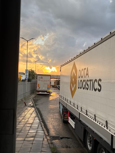 The image depicts an outdoor scene at a logistics or border crossing point in Kapitan Andreevo, Bulgaria, at either sunrise or sunset, characterized by a dramatic sky with golden light filtering through clouds, casting a warm glow on the wet ground.
In the foreground, a large white curtain-sided semi-trailer truck, identified by "DOTA LOGISTICS" in black lettering with an orange and yellow geometric logo on its side, occupies the right half of the frame. A "VOLVO" logo is visible on the rear section of the truck's chassis. The truck is positioned on a wet asphalt surface.
To the left, a paved sidewalk runs parallel to the roadway, bordered by a metal fence. The sidewalk is wet and highly reflective, showing grid patterns of light and shadow from the strong low sun. A red cylindrical object lies on its side on the sidewalk near the fence. Further down the street, beyond the fence, a white vertical sign displays "FRIGO" in blue lettering.
In the mid-ground, a second white semi-trailer truck is visible, displaying "NALÇACILAR" in red text and "SCHMITZ," along with Turkish contact numbers (e.g., beginning with "+90"). This truck's license plate appears to be Turkish ("34 AK 188"). Behind this truck, a sign clearly shows "БЪЛГАРИЯ" (Bulgaria) in Cyrillic, with "ОПЕРАТИВЕН" (Operational) partially visible beneath it, indicating a border control area. Multiple streetlights are visible throughout the scene. The sky features a mix of dark grey clouds and brightly illuminated, golden-toned clouds near the horizon. No people are visible.