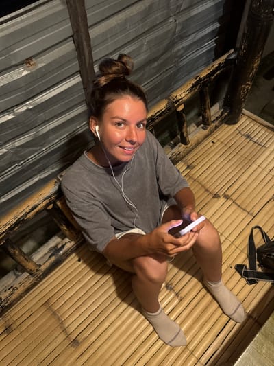 A woman is sitting on a bamboo-slatted floor, wearing a grey t-shirt, white shorts, and light-colored socks. She has her hair pulled up in a bun and is wearing white earbuds. In her hands, she is holding a white smartphone with its screen illuminated. The background consists of a corrugated metal wall and a wooden railing. A black bag is visible to the right of the woman. The setting appears to be an outdoor, rustic structure. The location is Kertasari, Indonesia.