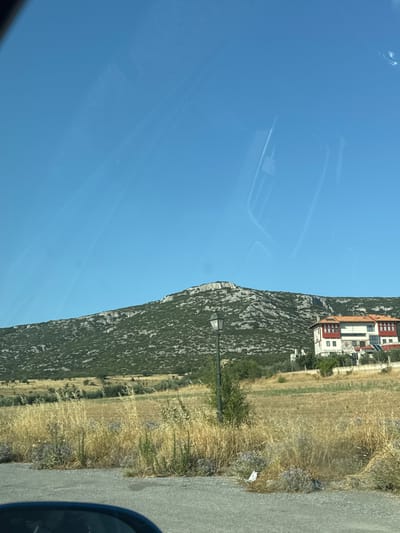 The image depicts a rural landscape under a clear blue sky. In the foreground, a grey asphalt road is visible, with the dark curved edge of what appears to be a vehicle's dashboard or interior in the lower left corner. Beyond the road, dry, golden grasses and scattered purple wildflowers dominate the middle ground. A traditional-looking street lamp stands prominently in this area. Further back, a verdant, rocky hillside rises, covered in low-lying green vegetation and sparse trees. On the right side of the hillside, a multi-story building with white walls, red trim, and a terracotta roof is situated. The building has multiple windows and balconies. The overall scene suggests a sunny day in a natural, possibly Mediterranean, setting. The specific location in Πετράλωνα, Greece, is not visually identifiable from the image itself. There are no people, animals, or overt actions depicted in the scene.