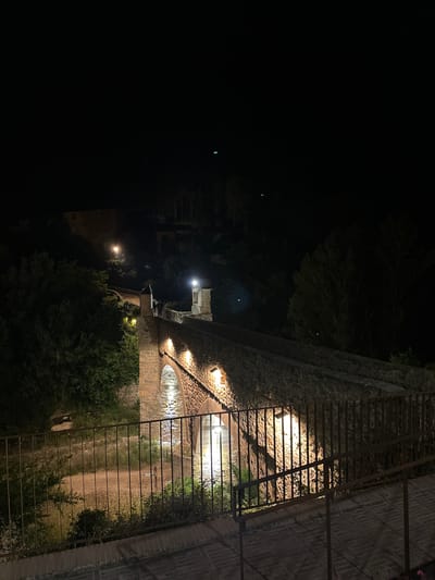 The image shows a night view in Montanejos, Spain. A stone aqueduct is illuminated by lights that shine from underneath the stone arches. A metal fence runs along the aqueduct on the right. Trees and foliage are visible in the background. The foreground is dark, with some illuminated structures visible in the distance. The aqueduct is the primary subject, with the fence, lights, and surrounding vegetation contributing to the overall scene.