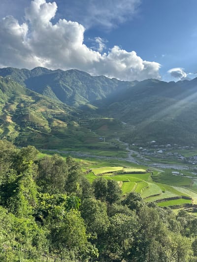A high-angle, wide shot captures a verdant mountainous valley in Tú Lệ, Vietnam, under a partly cloudy sky. The upper portion of the frame features a bright blue sky partially covered by large, white and grey cumulus clouds. Sun rays are visibly descending from the upper right, piercing through the atmospheric haze. The background is dominated by multiple layers of densely forested, dark green mountains, their slopes displaying some signs of agricultural terracing at higher elevations.

The midground reveals an extensive network of bright green rice terraces that conform to the natural contours of the valley walls and floor. A light-colored, rocky river or stream meanders through the center of the valley, separating sections of the terraced fields. Adjacent to the river, a compact village cluster is visible, composed of numerous houses with dark-colored roofs. Narrow paths or roads are discernible winding through the terraced landscape and connecting the village structures. The foreground is filled with a dense canopy of lush, dark green trees and foliage, indicating an elevated viewpoint overlooking the valley. No human figures or discernible activities are present in the scene.
