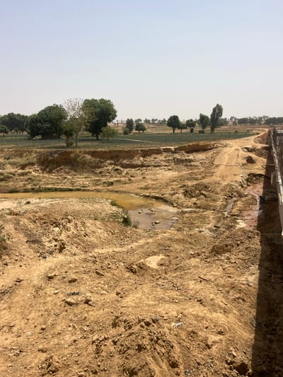 This image depicts an expansive outdoor scene in Wuciciri, Nigeria, observed under a clear, bright, and possibly hazy daylight sky. The foreground and midground are dominated by disturbed, dry, light brown earth, characterized by extensive erosion, dirt mounds, and visible tire tracks or paths.
In the mid-ground, an irregular body of turbid, brownish-yellow water is present, suggesting a shallow stream or large puddle. To its immediate right, a smaller, darker pool of water collects near a man-made structure. Beyond the immediate disturbed terrain, a flat expanse of cultivated green crops, appearing to be row-planted, is visible. This cultivated area is slightly elevated from the foreground by an eroded dirt embankment. Further into the background, numerous scattered trees with green foliage form a sparse tree line against the horizon.
On the far right side of the image, a partially constructed grey concrete structure is present, possibly a bridge abutment or culvert. It shows finished concrete walls and exposed elements that may be reinforcement bars or formwork. A dark blue object, resembling a tarp or fabric, is visible near the base of this structure. No individuals are present in the scene. Scattered small pieces of light-colored debris are visible on the disturbed ground in the foreground.