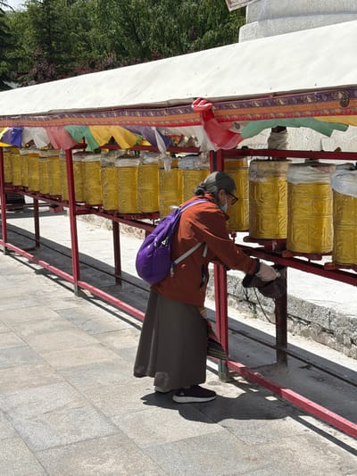 An elderly person is captured in an outdoor, eye-level shot, actively engaging with a row of prayer wheels under bright, clear sunlight. The subject, likely a woman, has grey hair pulled back and is dressed in an orange-brown jacket, a dark long skirt, and dark athletic shoes with white soles. She wears sunglasses, a light blue face mask, and white gloves on her hands, and carries a purple backpack over her left shoulder.
She is depicted turning one of the golden prayer wheels, which are intricately embossed with Tibetan script or symbols. These prayer wheels are systematically arranged in multiple rows within a sturdy red metal frame. Above the prayer wheels, a line of colorful prayer flags in red, yellow, green, white, and blue hangs down from a rich, patterned decorative strip that runs along the top of the structure.
The ground is paved with light-colored stone, and strong, defined shadows cast by the metal frame indicate direct and bright sunlight, suggesting it is midday or early afternoon. In the background to the left, a white wall-like structure and lush green trees are visible. The presence of numerous prayer wheels and prayer flags strongly identifies the location as a Tibetan Buddhist religious or cultural site, consistent with the given context of Chengguan District, China.