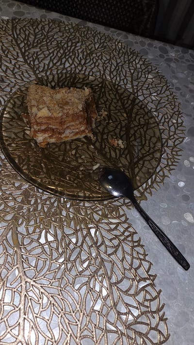A piece of cake sits on a brown glass plate, which is placed on a golden placemat with a leaf-like pattern. A dark spoon rests on the plate, next to the cake. The table appears to be covered with a white, patterned tablecloth. The background is out of focus and shows dark and striped patterns. The scene is set indoors, likely in a dining area. The image was taken in the city of Taezhny, Russia.