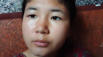 The image shows a close-up of a young person's face, with a focus on their eyes, nose, and mouth. The person has dark eyes and eyebrows, and their skin appears smooth with some subtle highlighting. They are wearing a garment with a red and gray pattern. The background is out of focus, with a textured wooden surface on the right and a patterned material on the left, possibly wallpaper or fabric. The lighting suggests an indoor environment. The image is a selfie, taken from a slightly upward angle. The location in Uchkun, Kyrgyzstan, is not directly indicated by any visual cues within the image itself.