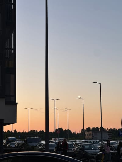 The image depicts a parking lot at dusk in Bugry, Russia, likely near the street Shosseynaya. A modern building is visible on the left, with several floors of dark-paneled windows. A tall, slender black lamppost dominates the center of the frame, extending from the bottom to the top. In the background, a line of trees forms the horizon. The sky transitions from a pale blue at the top to a warm orange and pink gradient near the horizon, indicating sunset. Multiple streetlights, some illuminated, are scattered across the scene. The parking lot is filled with various cars, including several vans and sedans. In the foreground, parts of a dark car are visible. Several individuals are present, some walking or standing among the vehicles. One woman in the middle ground is wearing a pink hoodie. The overall atmosphere is calm, with the scene illuminated by the fading daylight and artificial lights.