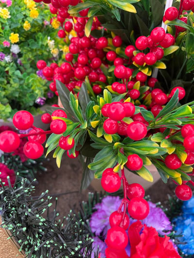 This close-up image features a vibrant display of artificial floral arrangements, with a dominant focus on glossy red berries and green foliage. Clusters of bright red, spherical berries, resembling cranberries or holly, are arranged on stems with green and yellow-green artificial leaves. The berries exhibit a distinct, reflective sheen.

In the background and foreground, other artificial flowers and decorative elements are visible. To the bottom left and center, smaller yellow, white, pink, and purple artificial flowers bloom amidst green foliage, some appearing slightly out of focus. To the right, larger, ruffled artificial flowers in shades of red, blue, and purple are present. A unique dark, spiky material with light green tips, possibly part of a wreath or base, is also seen on the right side.

The scene appears to be a still life display of decorative items, likely in a retail environment such as a craft store, flower shop, or market. The lighting is bright and even, highlighting the artificial textures and vivid colors of the plants. There is no visible activity or text. The overall impression is one of artificial decor, meticulously crafted for ornamental purposes.
