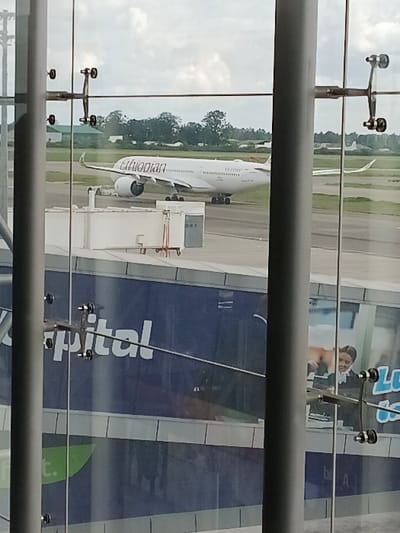 The scene depicts an outdoor view from within a modern airport terminal at Robert Gabriel Mugabe International Airport in Harare, Zimbabwe. The foreground is defined by two grey structural pillars and large glass panels secured by metallic spider fittings. These glass panels reflect a blue advertisement displaying the partial word "pital" and a female figure in uniform. Through the glass, a white Ethiopian Airlines Airbus A350 aircraft, identified by its livery and registration ET-AVF on the fuselage, is stationary on a dark tarmac surface. A white mobile passenger boarding bridge is visible on the tarmac to the left of the aircraft's nose. The background consists of a grassy field, numerous green trees, and distant low-rise buildings under a cloudy sky. No individuals are explicitly visible in the external airport environment.