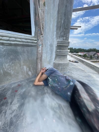 An elevated image captures a single individual lying prone on a grey concrete rooftop surface in Mns Asan, Indonesia. The person, oriented with their head toward the upper left, wears a dark t-shirt heavily stained with various paint colors and dark pants that exhibit a pronounced streaked or blurred artifact, particularly on the lower half. Their arms are bent, with hands covering their face. To the immediate left, an unpainted concrete wall with a horizontal molded design features an open window frame, revealing a dark interior. A rough wooden plank leans against this wall. Adjacent to the individual on the right, a vertical concrete column with multiple circumferential moldings rises. Exposed wooden construction beams are visible extending skyward from the right side of the structure. In the background, under a clear blue sky with scattered white clouds, a residential area with red-roofed houses and extensive green palm tree foliage is visible, extending towards a faint distant horizon suggesting a body of water.
