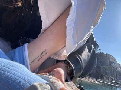 A close-up shot taken in Sorrento, Italy, shows a person with dark hair. The individual has an arm visible, bearing a tattoo of an abstract design. They are wearing a light-colored button-down shirt, a blue knitted sweater, and a black leather jacket. A variety of bracelets and rings adorn their hand. In the blurred background, there is a waterfront view with buildings. The sky is clear and blue, suggesting a sunny day.