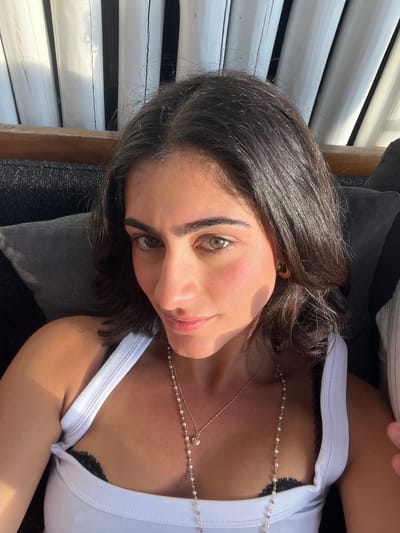 A woman with dark hair and green eyes lies on a dark grey patterned cushion. She wears a white tank top, under which a black lace bra is visible, and two gold necklaces. Her face is turned slightly towards the camera. Behind her are white vertical wooden slats, suggesting a shaded outdoor area. Based on the context of Cap-d'Ail, France, the location is likely a terrace or patio of a house or establishment with a view towards the sea.