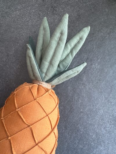 An overhead shot shows a stuffed orange carrot-shaped pillow with green fabric leaves on a dark gray textured surface. The carrot portion of the pillow is a textured orange fabric with crisscrossing lines of thicker fabric stitching to mimic the segments of a carrot. The green fabric leaves are stitched together and tied to the top of the carrot with a rough twine. The background is a dark, mottled gray surface. The location is not specified in the image.