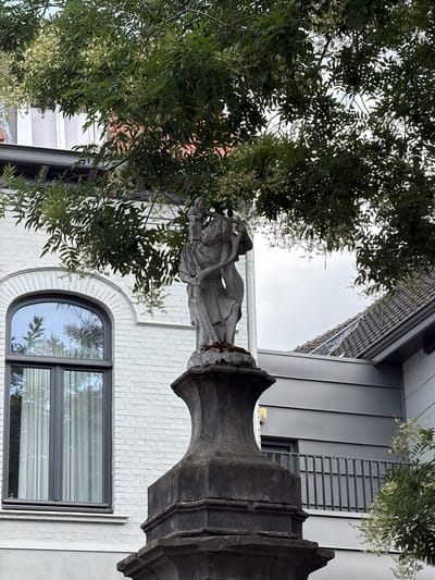 This image depicts a statue of two figures embracing, situated atop a stone pedestal. The statue and pedestal are made of weathered stone, showing signs of age and moss growth. Behind the statue, a white brick building with an arched window is visible. The window has a dark frame and sheer curtains. A tree with green leaves and small white flowers partially obscures the statue and the building. To the right of the statue, a modern building with grey siding and a metal railing is present, with a tiled roof extending into the background. The sky is overcast. The image appears to be taken from a low angle, looking upwards towards the statue and the tree. This scene is located in Roermond, Netherlands, specifically at the Roermond Municipal Museum.