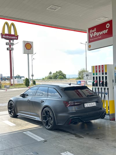 An Audi RS6 Avant, matte gray, is parked at a Shell gas station in Drazhevo, Bulgaria. The car, with license plate CB 0606 KO, is facing away from the camera, positioned at a gas pump. Overhead, a red and white Shell V-Power sign is visible, along with a McDonald's sign and pole in the background. The ground is a concrete surface with white markings. The sky is partly cloudy.