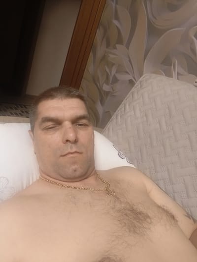 A man, shirtless and wearing a gold chain with a cross pendant, reclines on a bed. He is lying on his back, with his head resting on a white pillow patterned with a butterfly. His eyes are partially closed, and he has a neutral expression. The background features wallpaper with a floral design and a section of wooden paneling. The image appears to be a selfie, taken from a slightly elevated angle. The context of Petrovsk, Russia, is not visually discernible from the image itself.