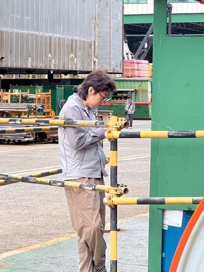 An outdoor, daytime, medium shot shows a person wearing glasses, a gray jacket, and tan pants standing in front of a yellow and black striped barrier. The person is looking down and appears to be holding a mobile device. In the background, there is a large industrial structure with corrugated metal siding and scaffolding. To the right of the main subject, another person in a gray shirt and dark pants walks away from the camera. A large green structure is visible on the right side of the frame. The scene appears to be in an industrial area in Geoje-si, South Korea, possibly a shipyard or manufacturing facility, given the presence of large industrial components and construction elements. The ground is paved with yellow and green markings.