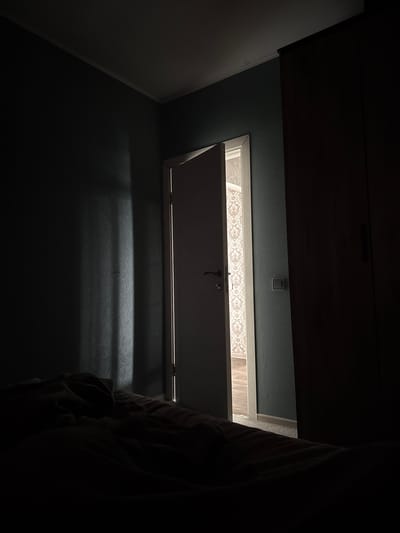 The image depicts an interior scene within a room, likely a bedroom, characterized by low lighting and dramatic shadows. A partially open door reveals a glimpse into an adjacent room with patterned wallpaper. The foreground is dominated by a dark, rumpled bed with blankets. The walls are a dark, muted color, and a vertical beam of light penetrates the darkness from the partially open doorway, casting shadows on the wall. A wardrobe or large piece of furniture is visible to the right, largely obscured by darkness. The environment suggests a dimly lit or nighttime setting. There are no visible people or animals. The location is identified as a room in Kondopoga, Russia.
