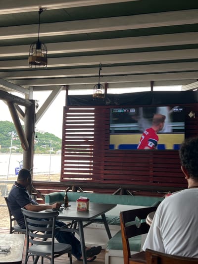 The image depicts a casual outdoor dining or bar area, possibly a beachside establishment, in San Juan del Sur, Nicaragua. In the foreground, a man wearing a dark polo shirt and jeans is seated at a table, appearing to be engaged with his phone. On the table are a beer bottle, a box of what appears to be beer coasters, and sunglasses. To the right, the back of another person wearing a white t-shirt is visible, also facing the television.
A large flat-screen television is mounted on a slatted wooden wall, displaying a soccer match with the score "BRA 0 - 0 NOR" and the time "25:15". The broadcast is from "26 FOX LIVE-NY NJ". The ceiling of the structure is supported by exposed wooden beams, and hanging from the ceiling are two industrial-style pendant lights with wire cages.
The background reveals a view of a beach, with the ocean and a small town or resort area situated on a hilly landscape. The overall ambiance suggests a relaxed setting, likely a place where patrons gather to watch sports and enjoy beverages by the coast.