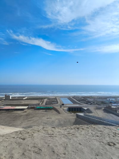 The image captures a high-angle view of Huanchaco, Peru, looking towards the Pacific Ocean. The sky is bright blue with scattered white clouds. A single bird is visible in flight. In the midground, there's a mixture of sandy terrain and low-rise buildings. A concrete canal and a road with a small bridge cut across the landscape. The foreground is dominated by a sandy dune, showing textures and patterns of the sand. The ocean horizon is hazy, with waves visible along the shoreline. No people are identifiable.