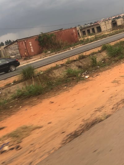 A road runs diagonally across the frame, bordered by a dusty, red-earth shoulder. Green vegetation grows along the road's edge. A shipping container and a partially visible black car are on the left. In the background, a grey sky looms over an unfinished building, presenting an overall slightly tilted perspective. Some scattered trash is also present near the vegetation.